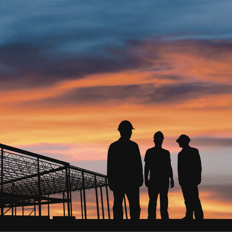 Siluetten av tre anleggsarbeidere foran en stålhall i solnedgang.